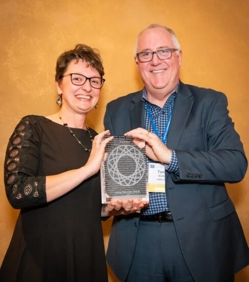 Two people smiling and holding an award together in front of a gold background.