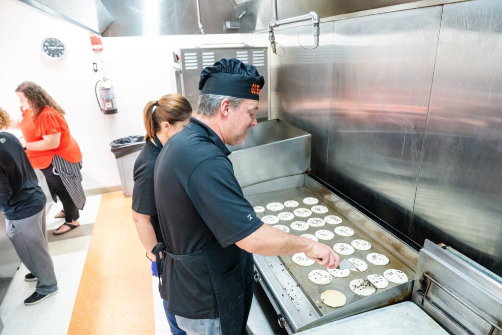 A man cooks multiple pancakes on a large griddle as others stand nearby in a commercial kitchen.
