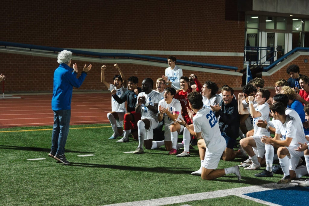 Soccer coach talks to excited players kneeling on the field at night near a track.