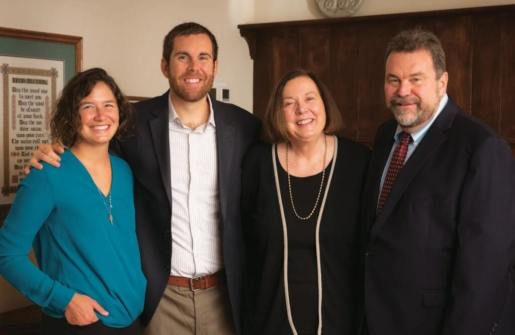 Four adults, dressed in business attire, smiling and standing together indoors.