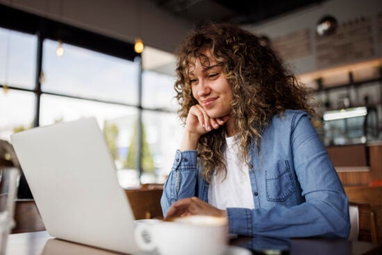 Smiling woman with curly hair uses a laptop at a café, with a coffee cup in the foreground.