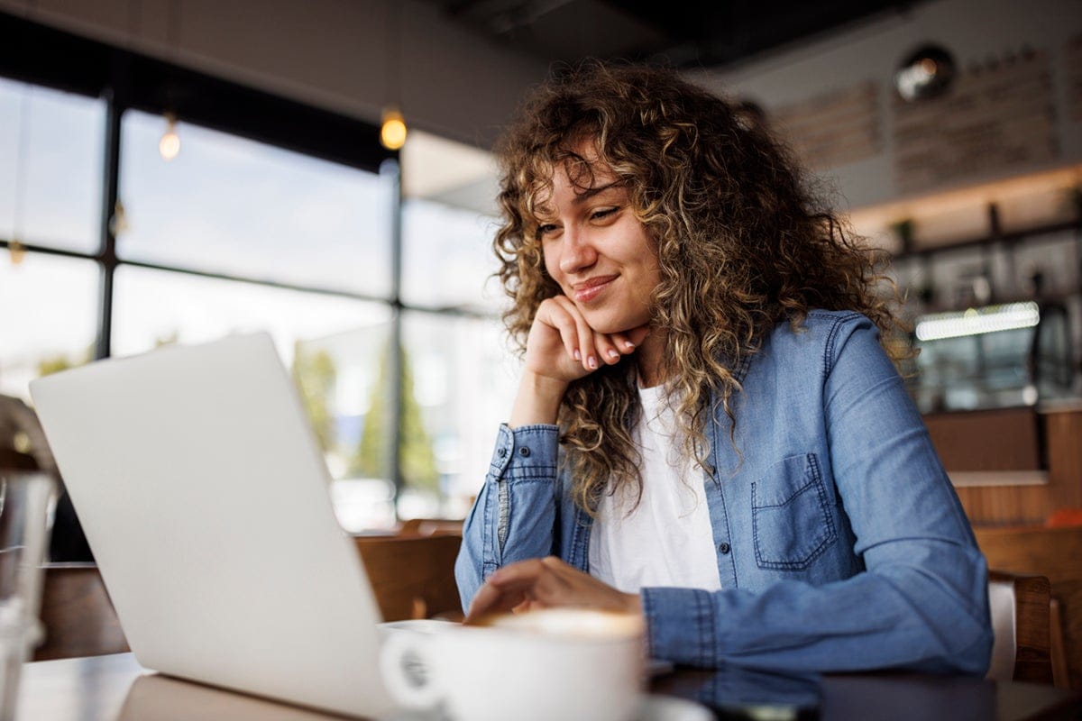 Smiling woman with curly hair uses a laptop at a café, with a coffee cup in the foreground.