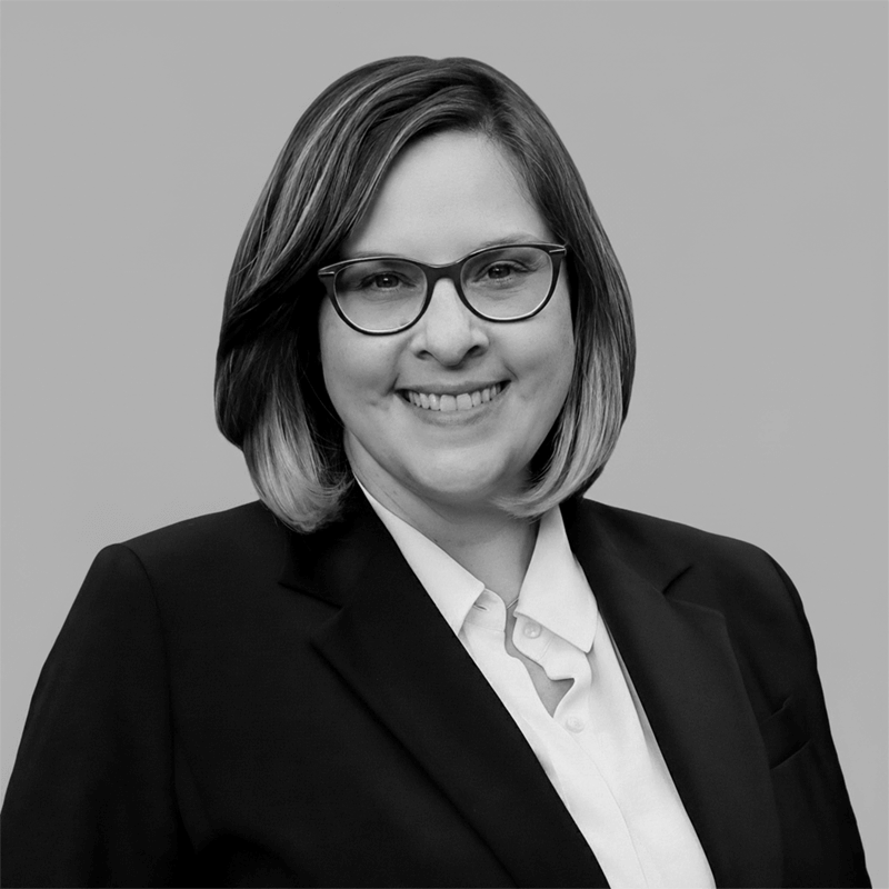 Smiling person with glasses and short hair, wearing a suit jacket and white shirt, on a plain background.