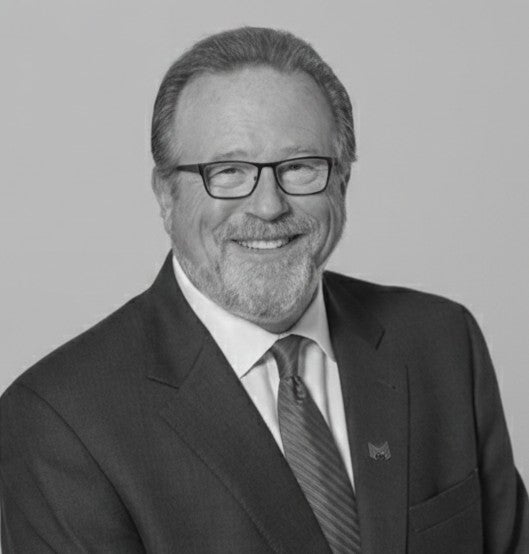 Smiling man with glasses and a beard wearing a suit and tie, posed against a plain background.