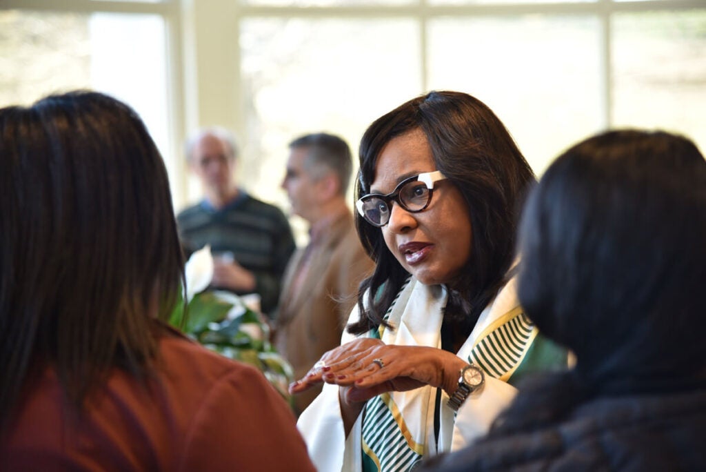 A woman in glasses speaks to two others indoors, with more people talking in the blurred background.