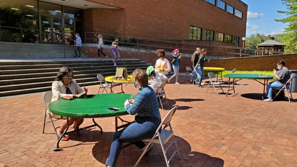 People sit at green tables outside a brick building, socially distanced and wearing masks.