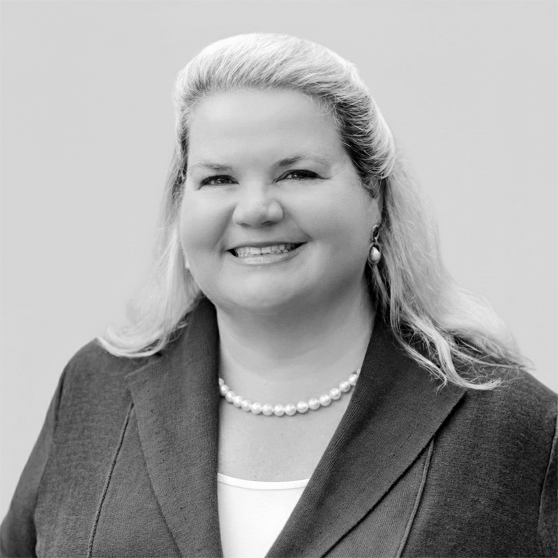 Smiling woman with long blonde hair, wearing a blazer, white top, pearls, and earrings, on a plain background.