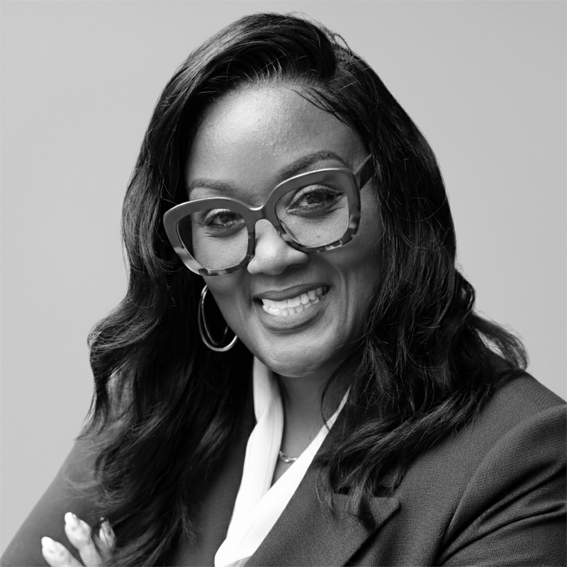 Smiling woman with glasses and wavy hair in a suit, posing confidently against a plain background.