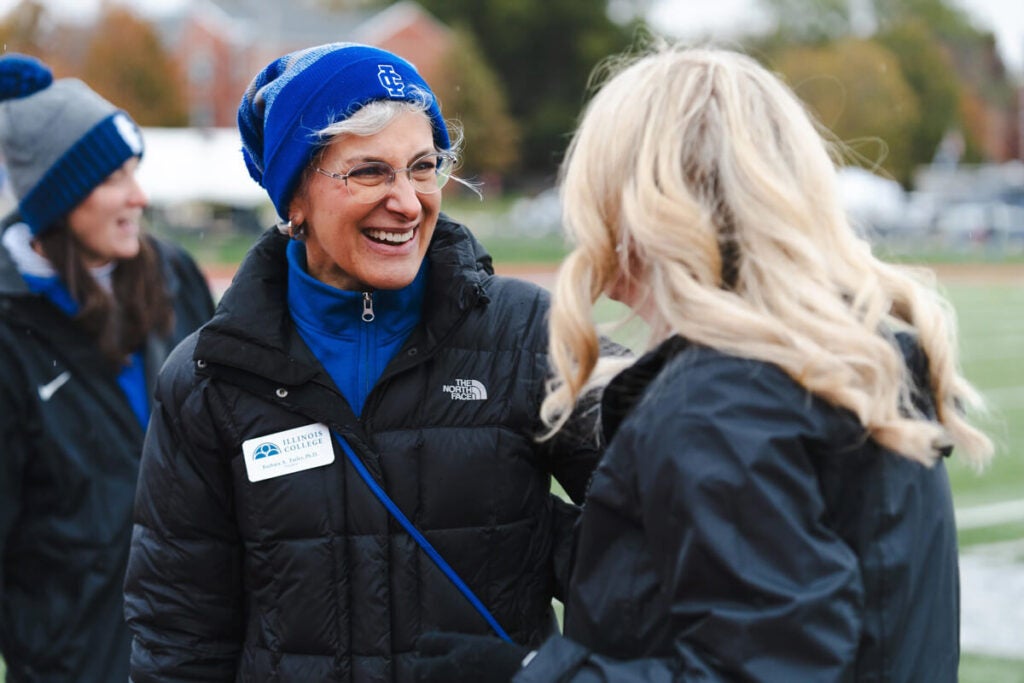 Two women in winter jackets smile and talk outdoors on a sports field, with another person in the background.