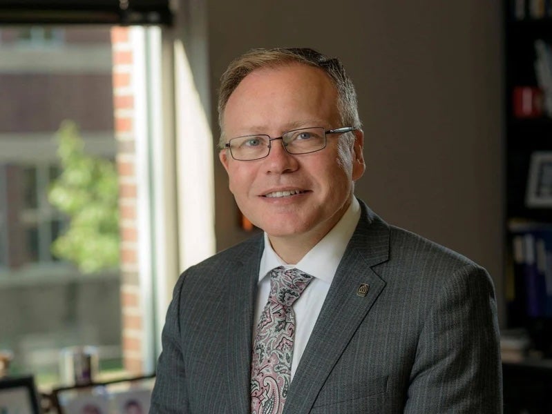 Smiling man in a suit and tie sits in an office with a window and shelves in the background.