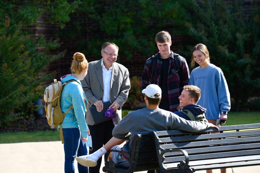 A group of five people, including a man in a blazer, talk and smile outdoors near a bench.