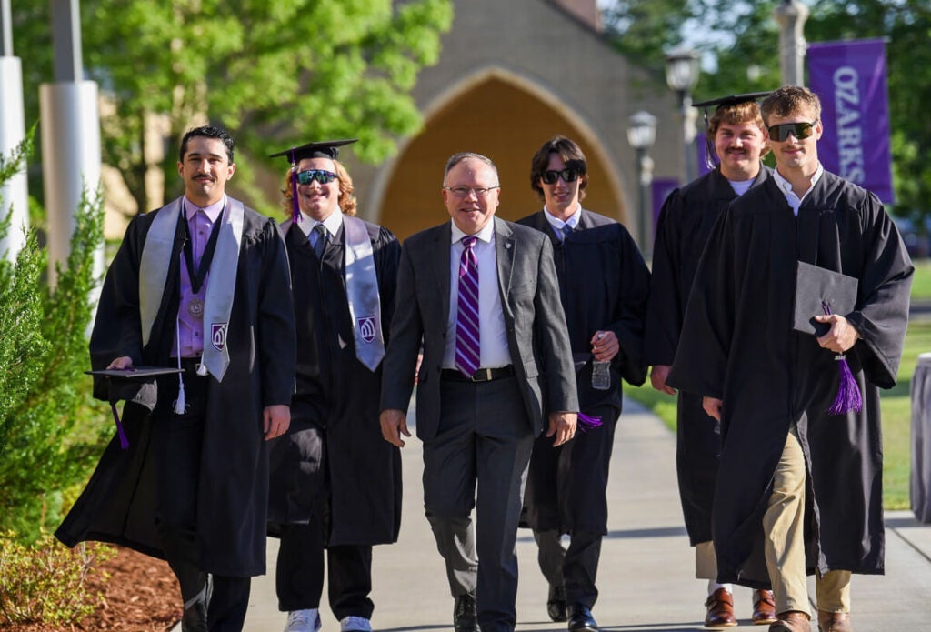 A group of graduates and a man in a suit walk together outdoors on a sunny day.