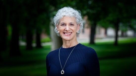 Smiling older woman with short curly gray hair, wearing a navy sweater and necklace, standing outdoors.