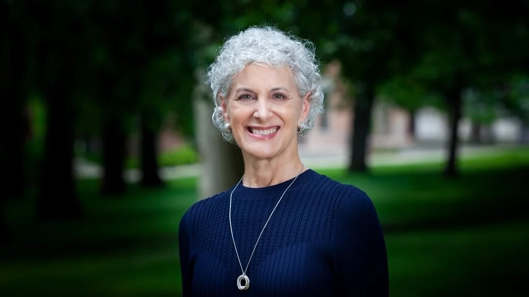 Smiling older woman with short curly gray hair, wearing a navy sweater and necklace, standing outdoors.