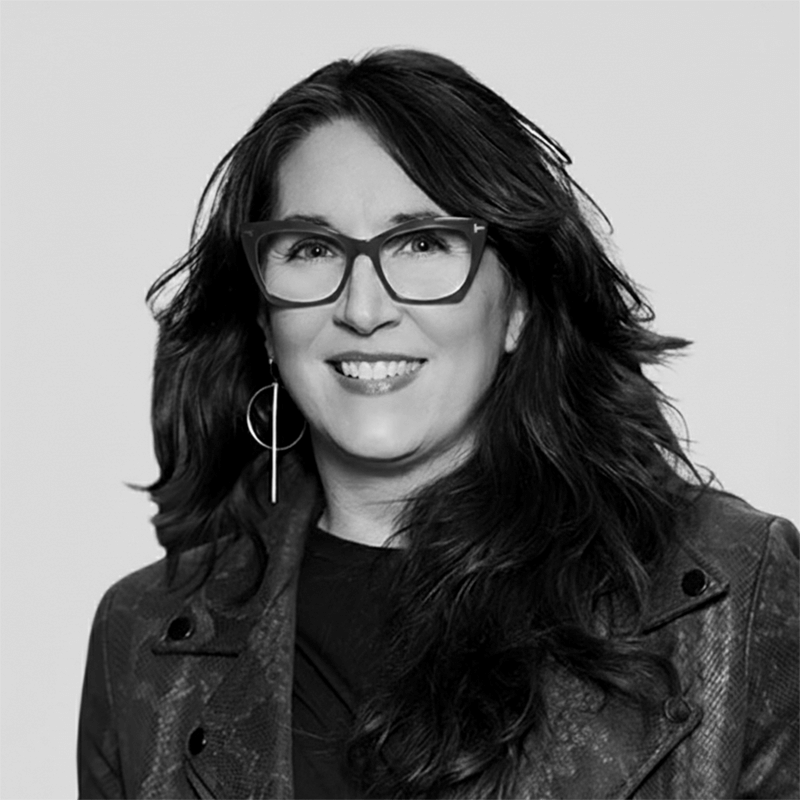 Woman with long dark hair, glasses, and large earrings, smiling in a patterned jacket against a plain background.