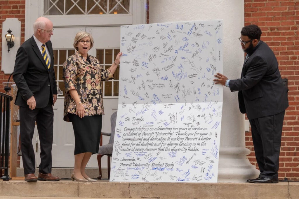 Three people stand outdoors holding a large card covered in signatures and a congratulatory message.
