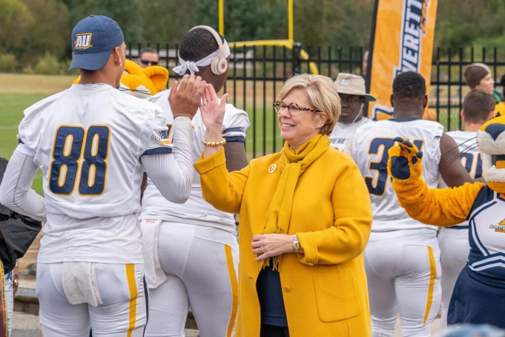 A woman in a yellow coat high-fives a football player on the sidelines during a game.
