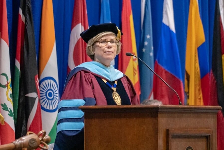 A person in academic regalia speaks at a podium with international flags displayed in the background.