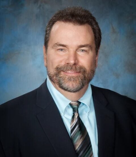 A man in a suit and striped tie smiles in front of a blue and gray studio background.