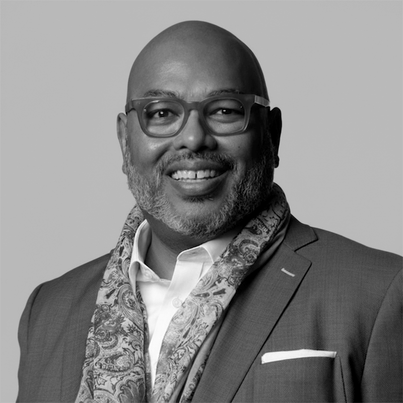 Smiling man in a suit, glasses, and patterned scarf, posed against a plain background.