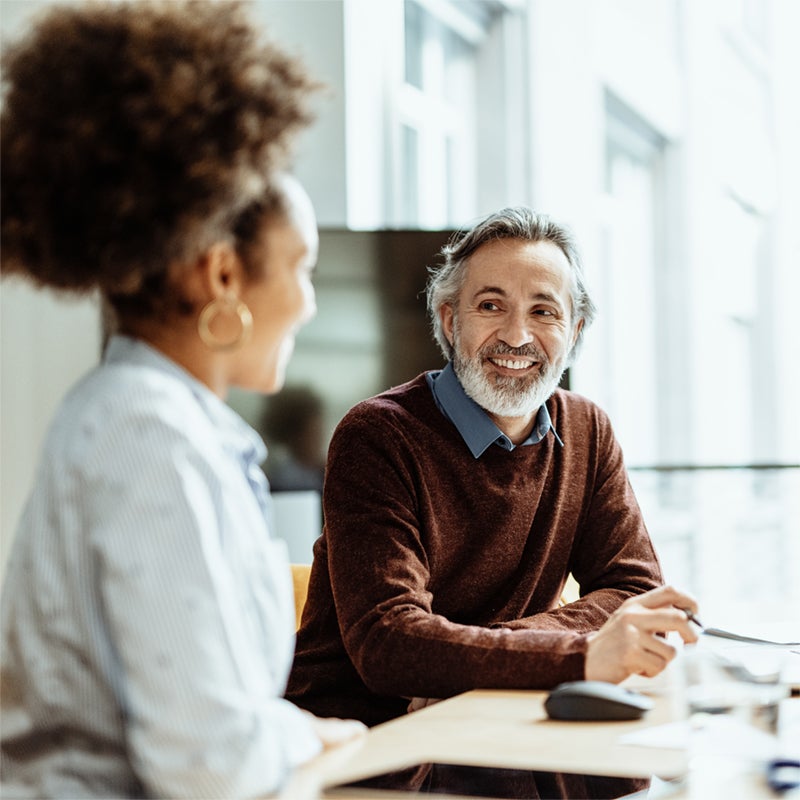 Man and woman sitting at a desk, smiling and talking in a bright office setting.