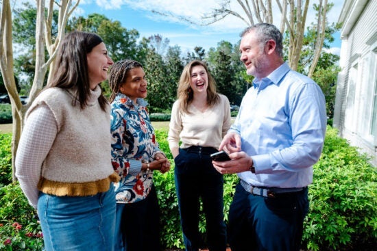 Four adults standing outside, smiling and talking together in front of green bushes and trees.
