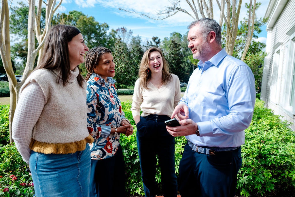 Four adults standing outside, smiling and talking together in front of green bushes and trees.