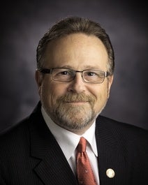 Man with glasses, beard, and mustache in a suit, red tie, and pin, smiling against a dark background.