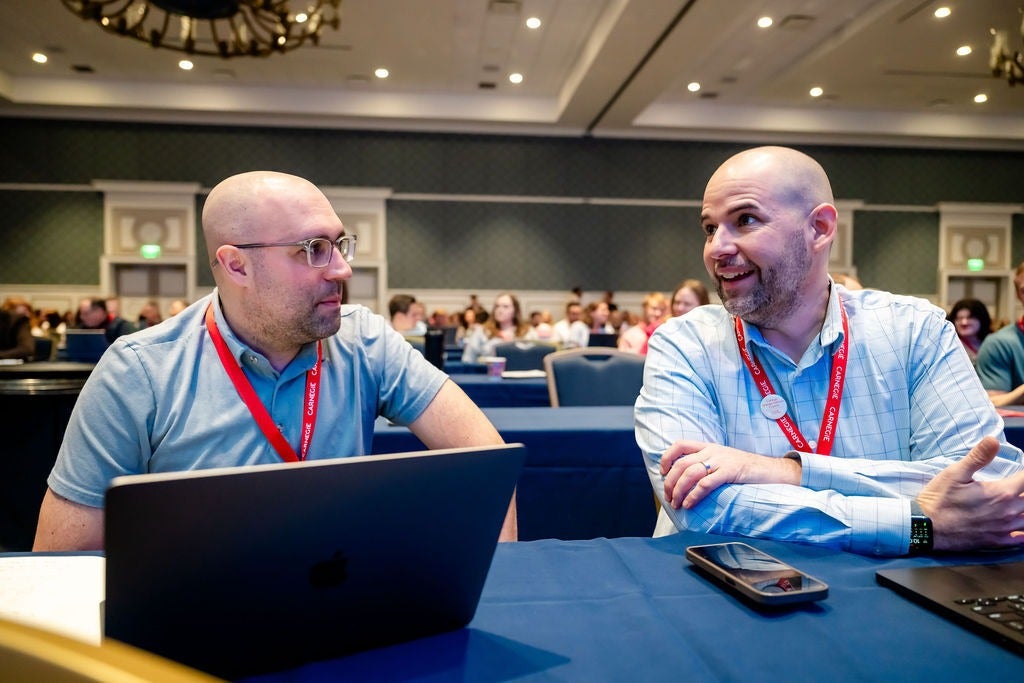 Two men with laptops and red lanyards talking at a conference table with people seated in the background.