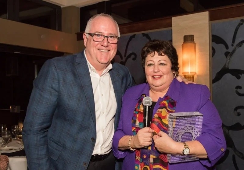 Two people smiling; the woman holding a microphone and a glass award, the man standing beside her.