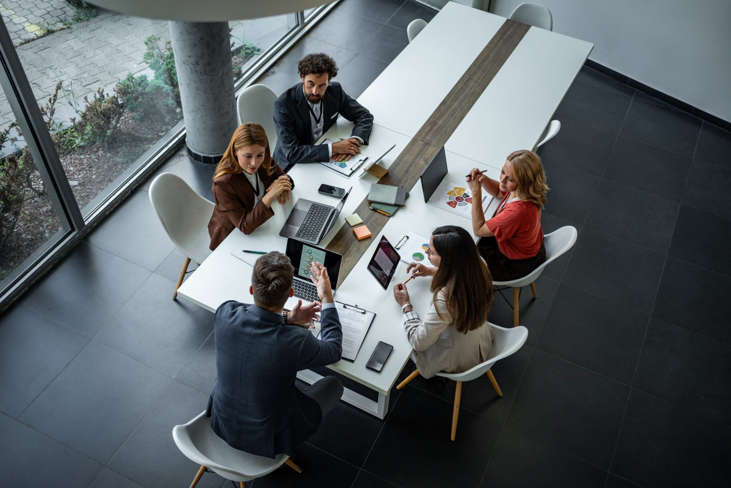 Five people in business attire sit around a conference table with laptops and papers, having a meeting.