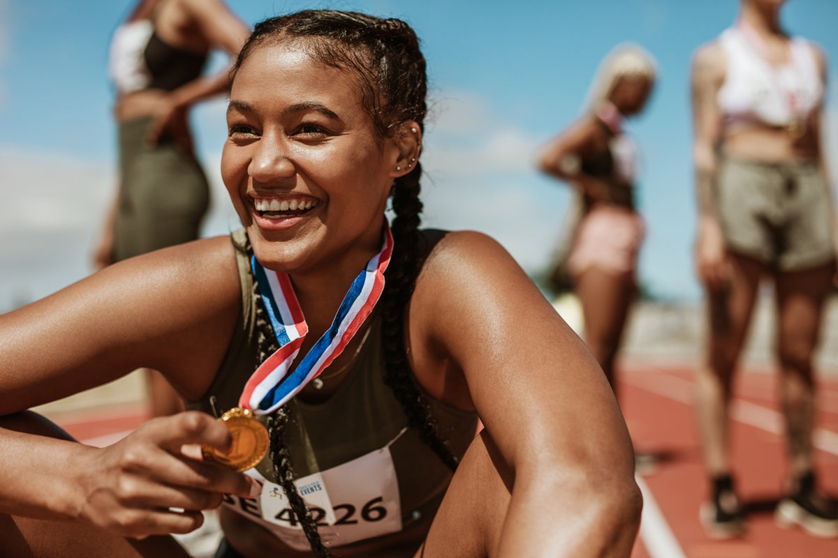 Smiling athlete with a gold medal sits on a track, teammates standing in the background.