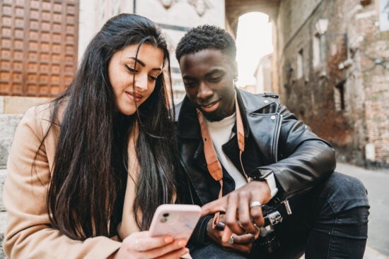 Two people sitting outside, smiling and looking at a smartphone together on a city street.