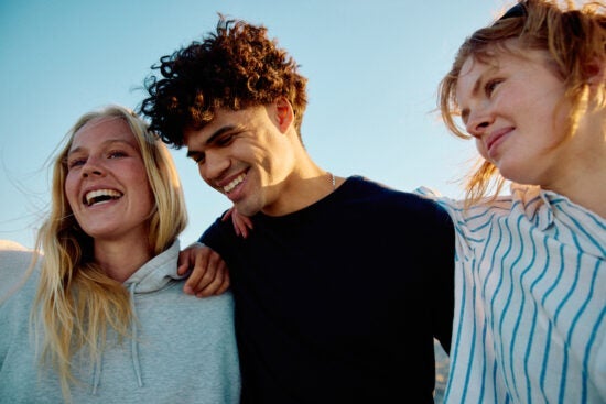 Three friends smiling with arms around each other outdoors under a clear blue sky.