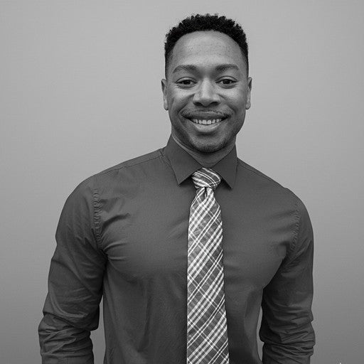 A man in a dress shirt and plaid tie smiles at the camera against a plain background.