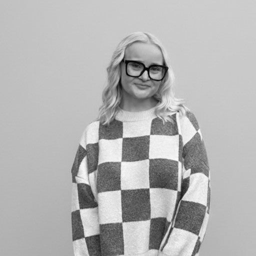 A woman with wavy hair and glasses, wearing a checkered sweater, smiles in front of a plain background.