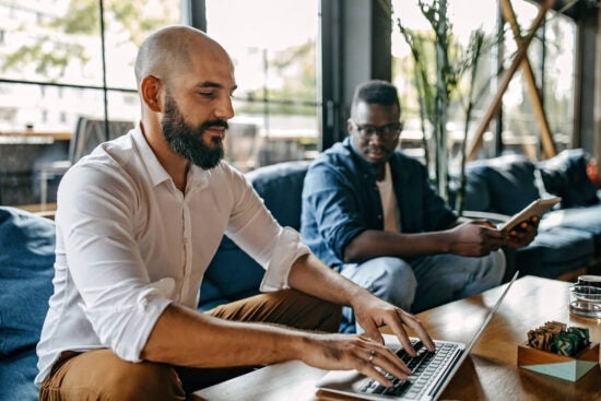 Two men sitting indoors; one types on a laptop while the other reads a book on a couch nearby.