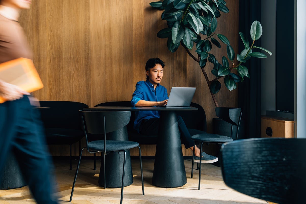 Man in blue shirt working on laptop at a modern cafe, with another person walking by in the foreground.