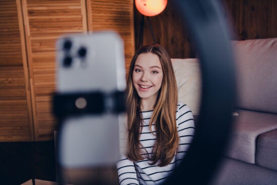 Young woman smiling at camera, filming herself with a smartphone and ring light in a cozy room.