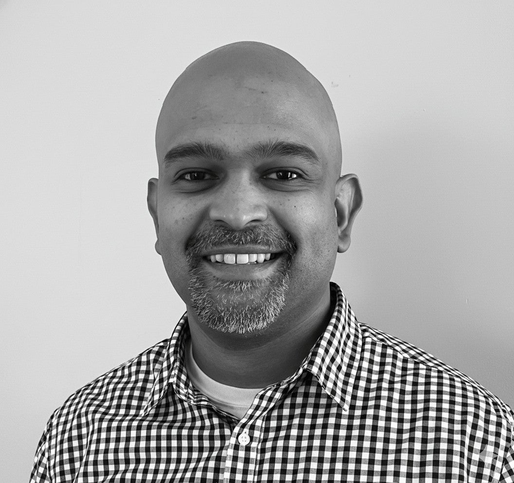 Smiling man with a shaved head and beard wearing a checkered shirt, standing in front of a plain background.