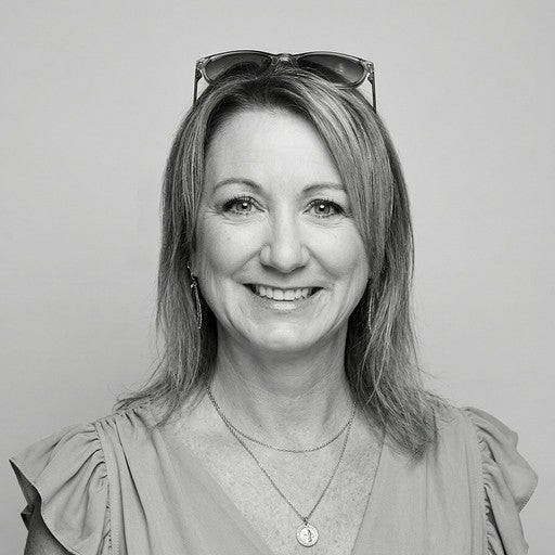 Smiling woman with shoulder-length hair, wearing glasses on her head and layered necklaces, in a light top.
