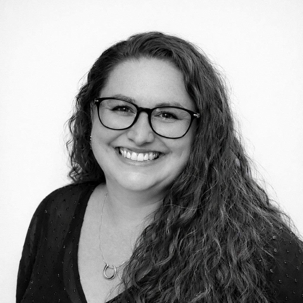 Smiling woman with long curly hair and glasses, wearing a dark top and necklace, in front of a plain background.