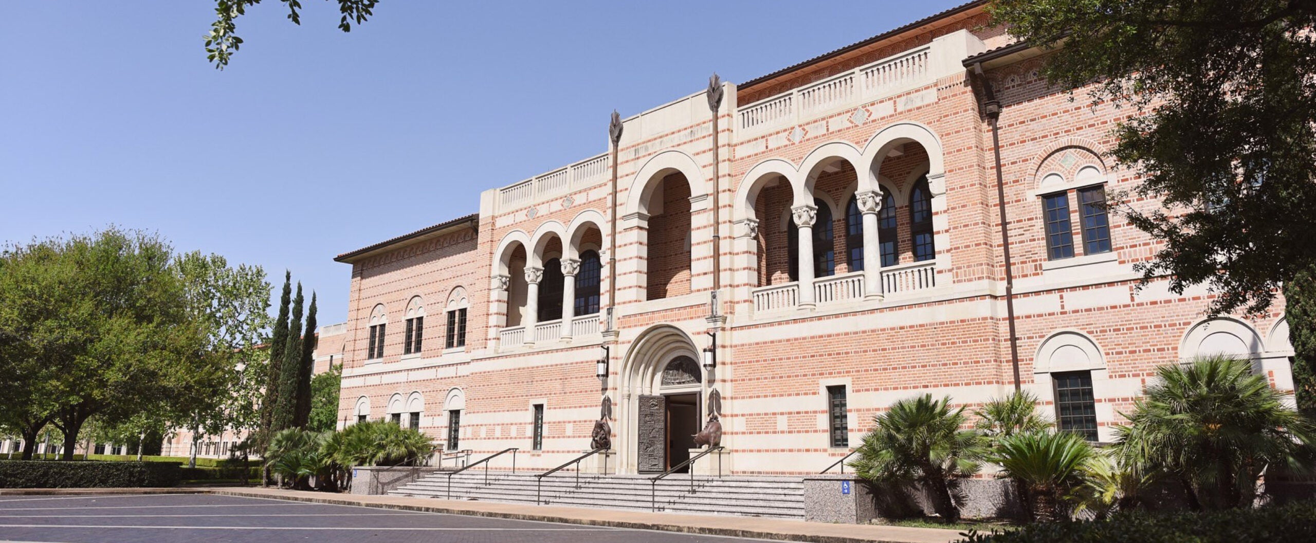 A large brick academic building with arched windows, palm trees, and steps leading to the entrance.