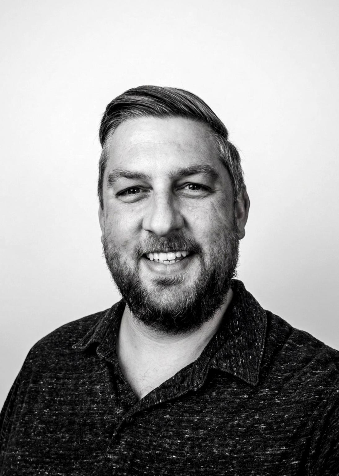 Smiling man with short hair and beard, wearing a dark collared shirt, in front of a plain light background.