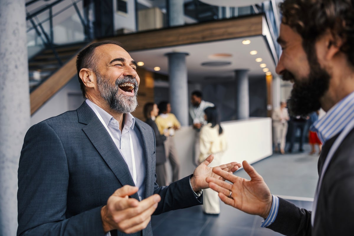 Two men in business suits laughing and talking in a modern office lobby with people in the background.