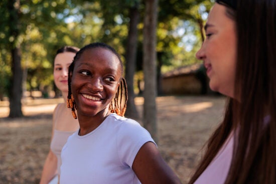 Three young women smiling and talking together outdoors in a park with trees in the background.