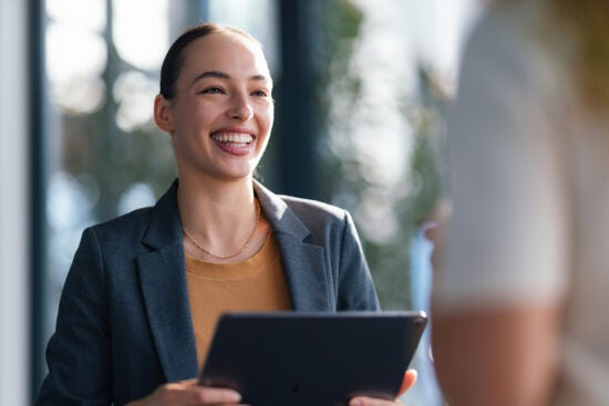 Smiling woman in a blazer holding a tablet, talking to another person in a bright office setting.