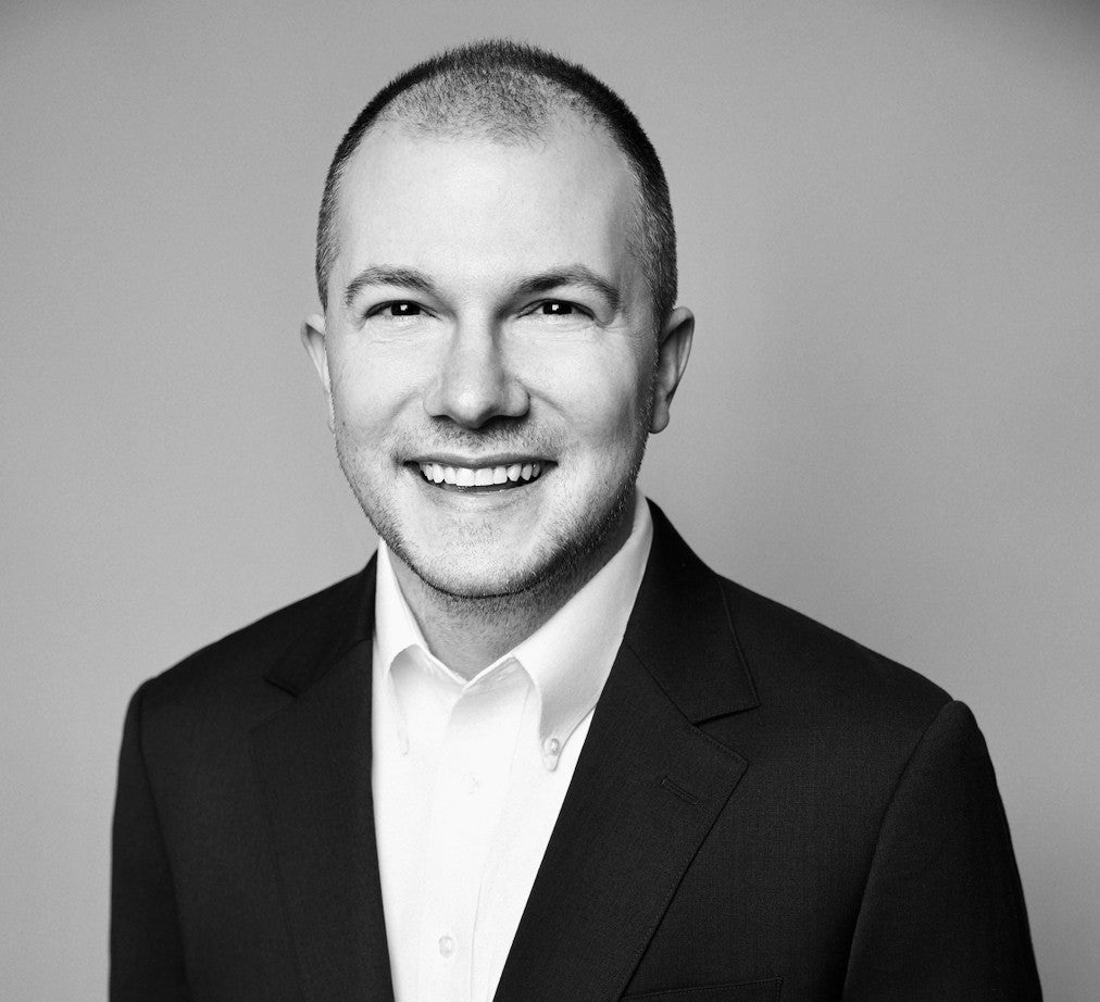 Smiling man in a suit jacket and white shirt against a plain background, black and white portrait.