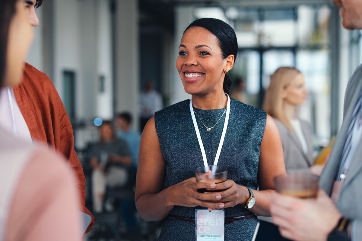 Smiling woman holding a drink and wearing a name badge at a social event, talking with others.