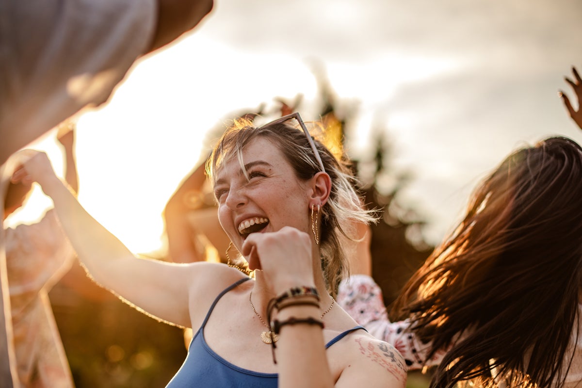 Young woman smiling and dancing outdoors with friends at sunset, sunlight glowing behind them.
